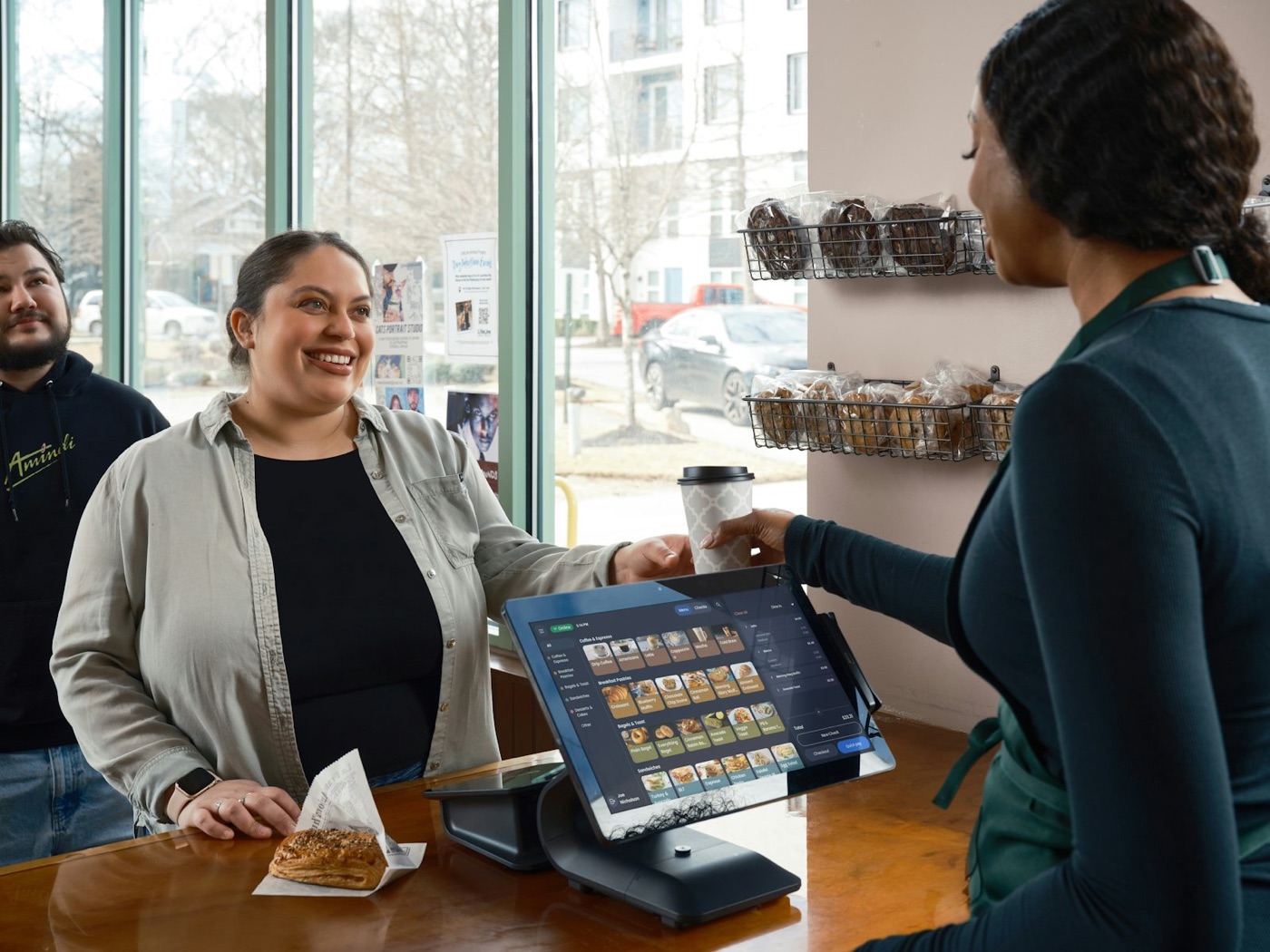 A barista hands a coffee to a smiling customer across a counter with a point-of-sale screen
